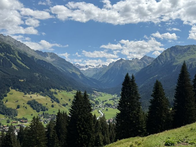 A panoramic view of a lush alpine valley nestled between towering, forested mountains under a partly cloudy sky, with a small village visible in the distance. The caption seems mismatched with the image, as this scene depicts a mountainous European-style landscape rather than Dubai's urban desert environment.