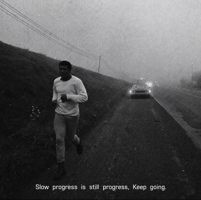 Black-and-white motivational image of a man jogging along a foggy roadside with a vintage car approaching in the distance, captioned 'Slow progress is still progress, Keep going.'