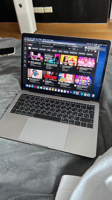 A MacBook Air laptop sits open on a gray blanket on a bed, displaying the YouTube website with various video thumbnails visible on the home page.