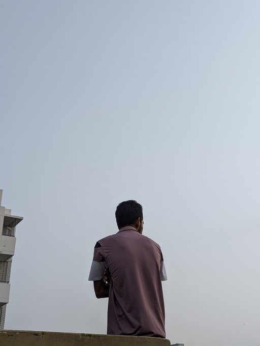 The image shows a man sitting alone on a ledge or wall with his back facing away from the camera. He appears to be wearing a purple shirt, which contrasts with the white building behind him. The sky above them has a hazy appearance, suggesting that they are in an urban setting. The post contains text about leveling up and learning, which could be related to the man's situation or the surrounding environment.