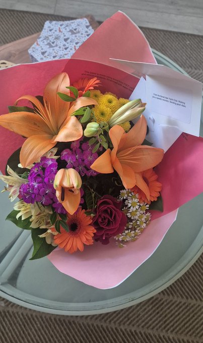 A vibrant bouquet of orange lilies, purple stock, red roses, and yellow accents wrapped in pink paper with a "from the team @ BoredApeYC, wishing you a speedy recovery" note sits on a mint green table, perfectly capturing the thoughtful BAYC get-well gift described in the post.