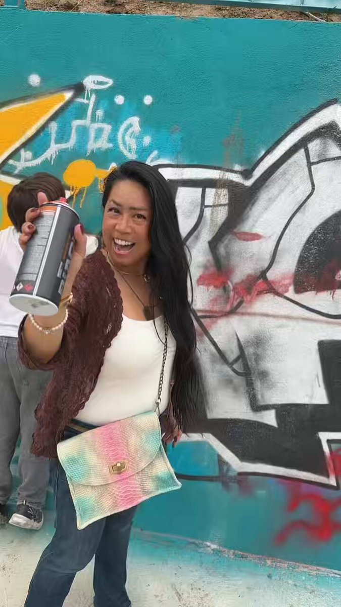 The image features a woman standing next to a graffiti-covered wall with her arms raised high. She is wearing a brown jacket and blue jeans, holding a spray paint can in one hand while smiling at the camera with the other. The background of the photo includes a green wall that has been transformed into an art piece by graffiti artists. There are also some people visible in the image, but they are not the main focus as the woman is the central figure.