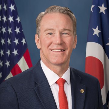 Senator Jon Husted of Ohio is shown smiling in a formal portrait, wearing a dark navy suit with a red tie, standing between an American flag and the Ohio state flag. The image accompanies news of his support for the CLARITY Act, reflecting his role as a U.S. Senator advocating for the legislation's passage.