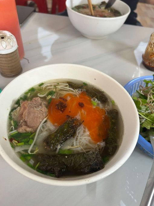 A bowl of Vietnamese-style noodle soup, likely bún, topped with bright orange chili sauce, meat, greens, and fried seaweed, served alongside a basket of fresh herbs and vegetables.
