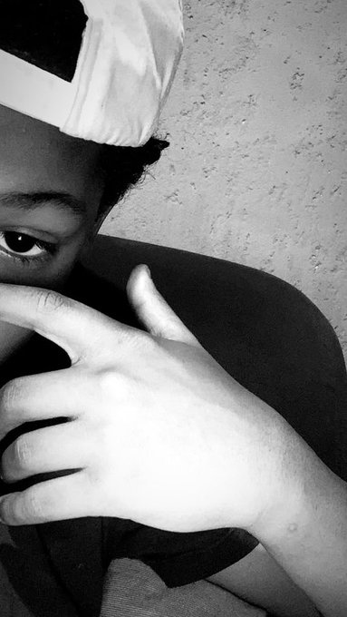 A close-up, black-and-white photograph of a child wearing a white hat, looking at the camera while holding a hand up near their face.
