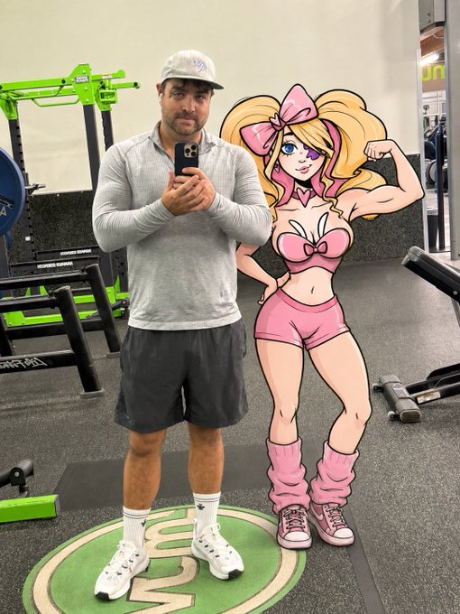 A man wearing a gray long-sleeve shirt, shorts, and a baseball cap takes a mirror selfie in a gym using his phone. Next to him is a large cutout of a cartoon character of a blonde woman in pink athletic wear flexing her bicep.