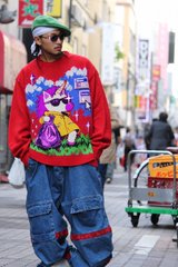 A person wearing a green hat, sunglasses, a red graphic sweater, and oversized blue cargo pants, standing on a street with shopping carts in the background.