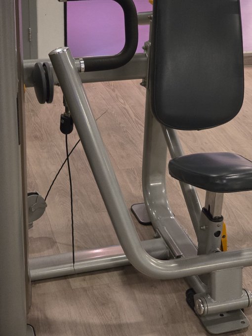 A close-up view of a silver and black gym weight machine in a fitness room, showing a cable that has snapped and is dangling loose. The broken cable visually humorously represents the frustration expressed in the caption about waiting for $XRP and $FLR to finally rise in value.