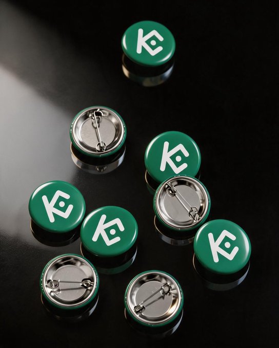 Green pin-back buttons featuring a stylized "KC" logo are scattered across a dark reflective surface, showing both their branded front faces and their metal clasp backs. The bold green-and-white design pairs perfectly with the post's invitation to pin them on a backpack, jacket, or trading cap.