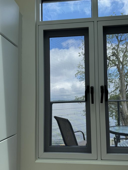 A view through a white-framed window showing a lake, sky with clouds, and a patio chair, taken from inside a room.