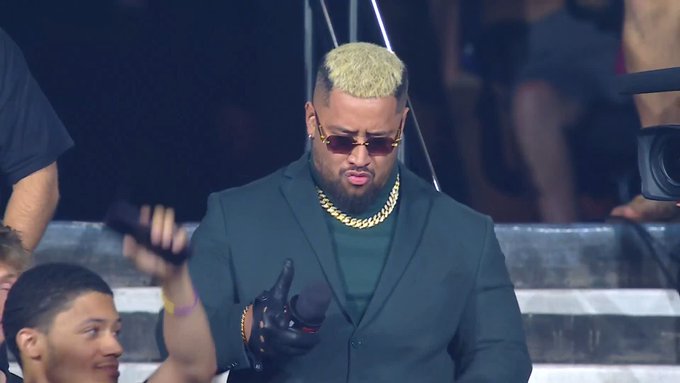 A man with a blonde buzz cut and beard, wearing sunglasses, a green suit, and a thick gold chain, holds a microphone while performing.