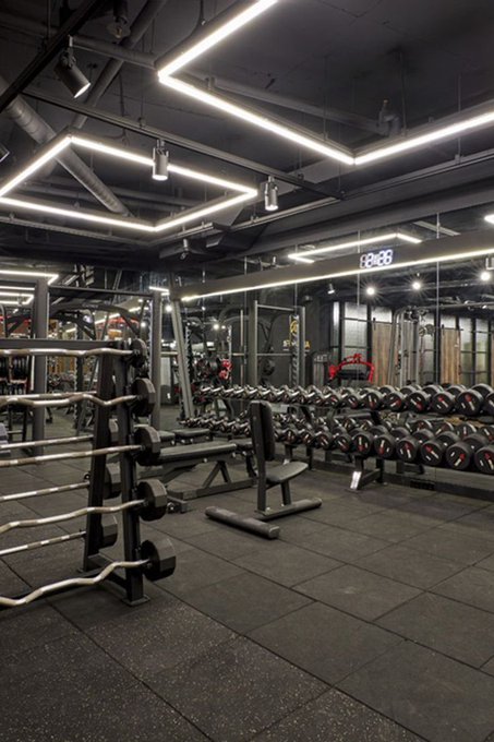 An empty, modern gym interior with black flooring, featuring a rack of dumbbells, a curl bar, benches, and distinctive square-shaped LED ceiling lights.