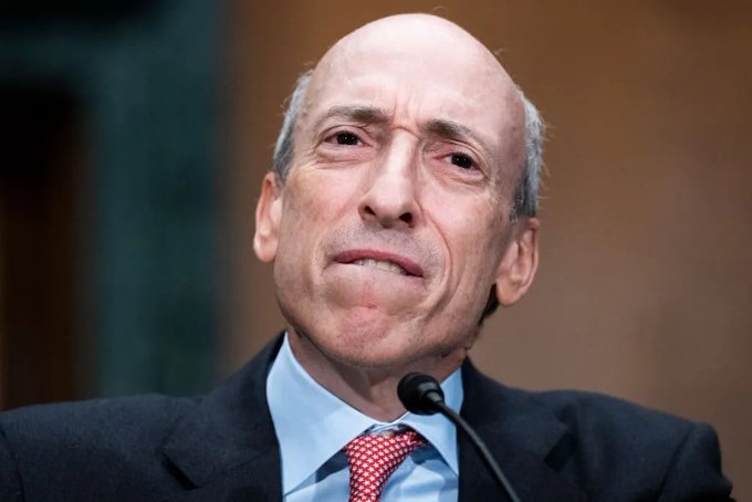 An older bald man with gray hair on the sides wears a dark suit and red patterned tie while speaking into a microphone.