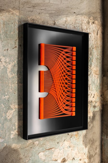 A framed piece of wall art featuring orange cables or tubes connecting two vertical bars, mounted on a textured stone wall.