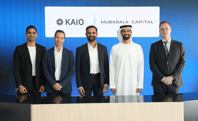 A corporate photo showing five men standing behind a counter; they are dressed in suits except for one in traditional white Arab attire, standing in front of a backdrop displaying the 'KAIO' and 'MUBADALA CAPITAL' logos.