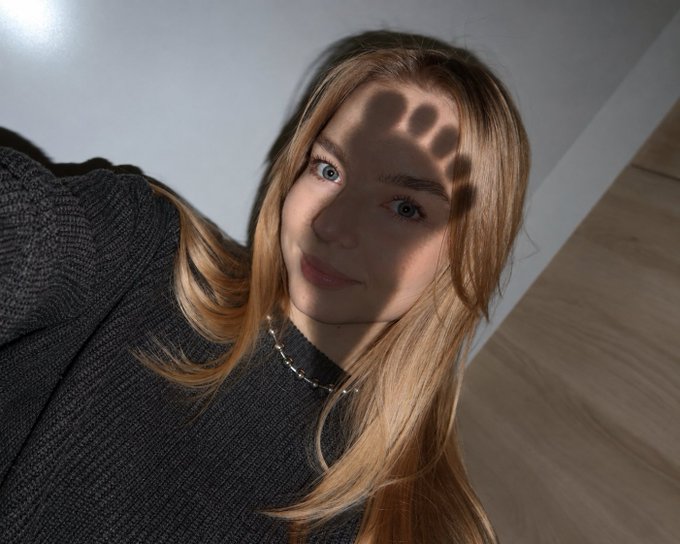 A close-up selfie of a young woman with long blonde hair wearing a dark gray sweater and a delicate necklace, with a hand shadow cast across her forehead.