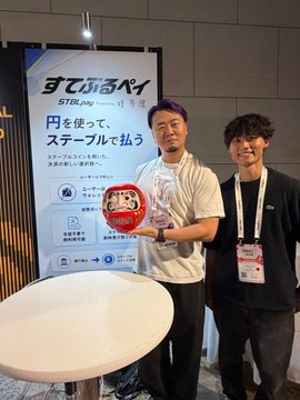Two men posing in front of a 'STeLPay' promotional banner with Japanese text 'すぐてるペイ' at what appears to be a trade show or expo booth.
