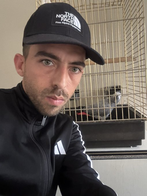 A selfie of a man with light eyes and stubble wearing a black The North Face cap and jacket, positioned in front of a large bird cage containing a dark-colored bird.