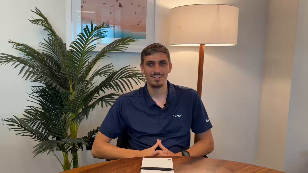 The image features a man sitting at a table, smiling for the camera while wearing a blue shirt. The setting appears to be an office or workspace with a lamp providing light over the scene. There are two potted plants visible in the room - one on the left side and another on the right side of the frame. A caption below the image reads "Tune into this CoFHE chat with our BD @l_woetzel and you might just catch some alpha on a potential confidential stablecoin".