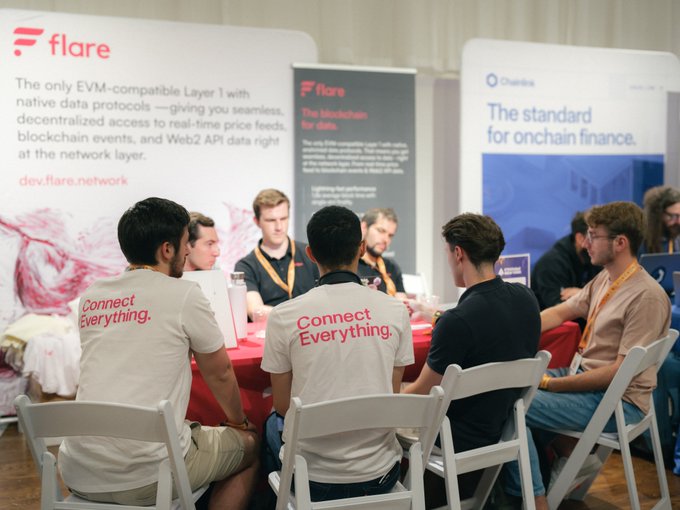 A group of conference attendees wearing branded "Connect Everything" t-shirts gather around a table at the Flare Network booth, which advertises itself as "The only EVM-compatible Layer 1 with native data protocols" (dev.flare.network), alongside a Chainlink banner promoting "The standard for onchain finance." The image reflects the weekend post's theme of community-driven building and experimentation in the blockchain space, capturing developers engaged in collaborative discussion at the event.