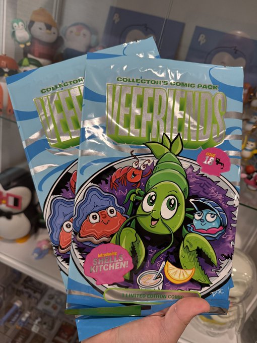 A hand holding two "VeeFriends" Collector's Comic Pack blind bags featuring colorful cartoon characters, marked as "Limited Edition Comics," inside what appears to be a collectibles store.