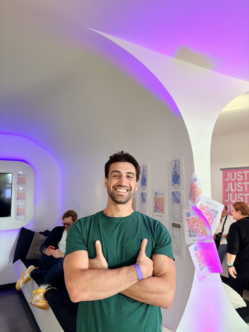 A smiling man in a green t-shirt gives a thumbs-up while standing inside a futuristic white pod-like structure with purple ambient lighting, with other people visible in the background.