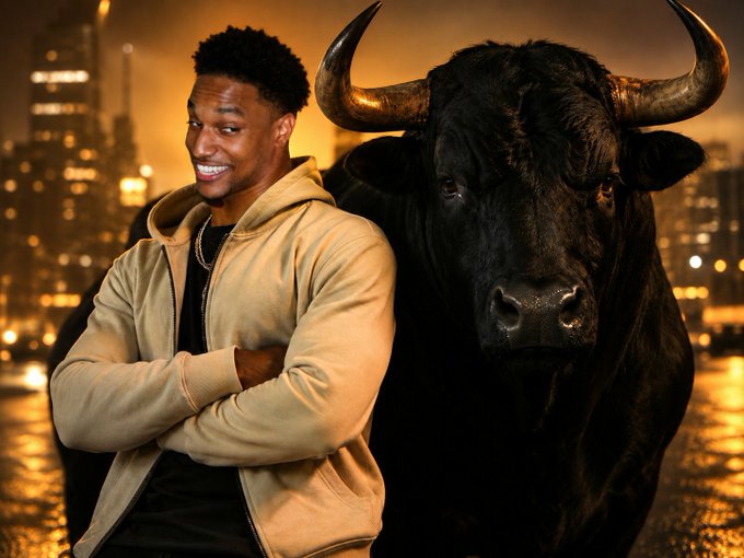 A smiling man in a tan hoodie stands with arms crossed next to a large black bull with prominent horns, set against a golden city skyline at sunset.