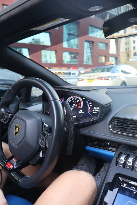 View from the driver's seat of a Lamborghini with the steering wheel and digital dashboard visible, parked on a city street with another luxury car seen through the windshield—capturing the aspirational "GM (get money)" lifestyle of high-end automotive luxury.