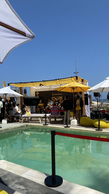 An outdoor Binance event (#Binance) is set up at a poolside venue with a yellow Binance-branded canopy, white umbrellas, bean bags, and attendees gathered on a wooden deck beside a swimming pool.