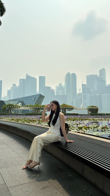 A woman in white pants and a white top sits on a wooden bench holding sunglasses, with the Singapore city skyline and Marina Bay visible in the hazy background.