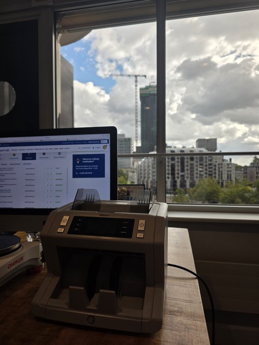 A currency counting machine sits on a desk in front of a computer screen displaying an OHS APP login page, with a city skyline visible through the window behind it—visually representing the cash proceeds being converted from silver sales into XRP and FLR crypto investments.