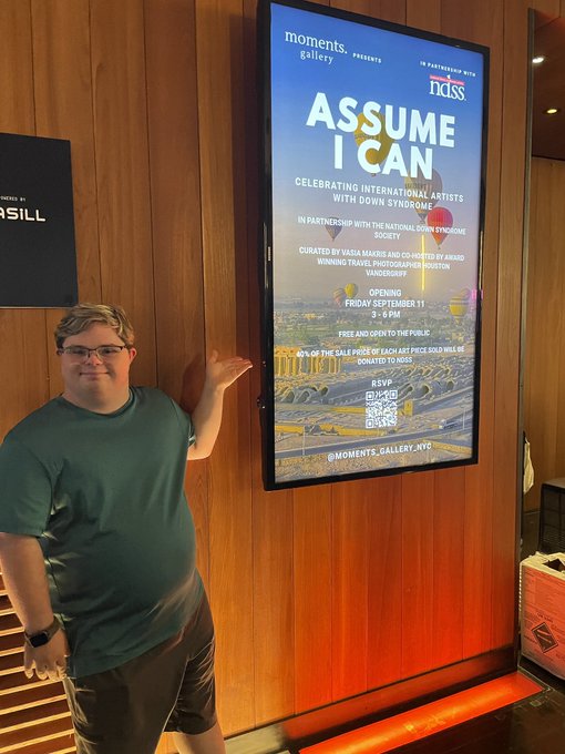 A man in a green t-shirt and glasses gesturing toward a digital poster that reads "ASSUME I CAN" for an exhibition at Moments Gallery NYC celebrating artists with Down Syndrome, on Friday September 11.