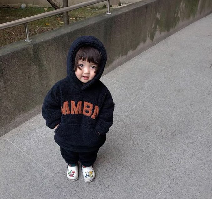 The image features a young child wearing a hooded sweatshirt with the word "mamba" printed on it. The child appears to be standing alone, possibly waiting for someone or just taking a break from playing outside. The background of the photo shows a sidewalk and a wall, providing context about where the child is located.