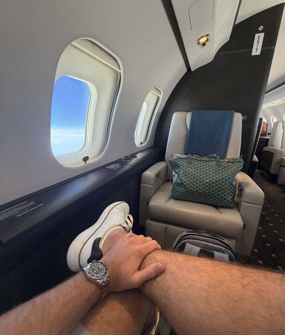 A man relaxes with his leg crossed in a spacious private jet cabin, wearing white sneakers and a luxury watch, with a green tote bag placed on the cream leather seat beside the window. The "EXIT" sign is visible overhead, aligning with the post's caption about enjoying a private jet experience.