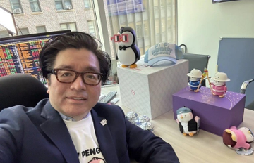 A man wearing glasses and a suit jacket over a 'PENGU' t-shirt sits at a desk featuring two computer monitors displaying stock charts, with several penguin plush toys displayed on pedestals beside him.