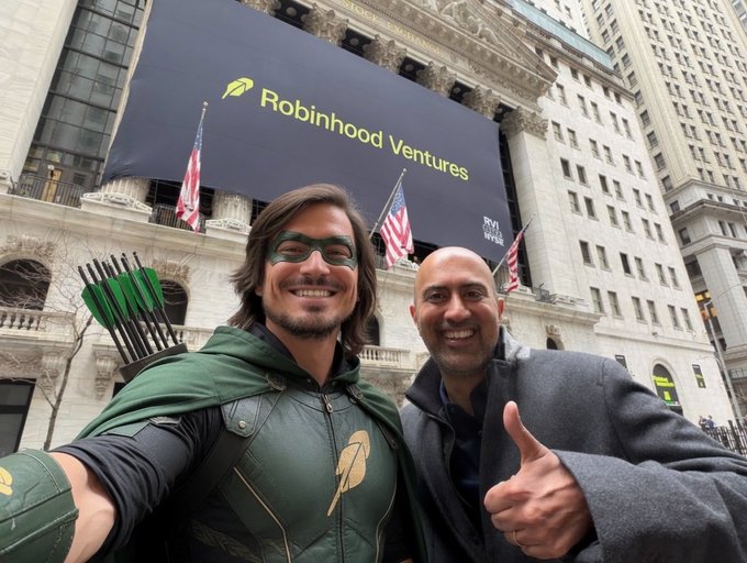 Two men pose outside the New York Stock Exchange beneath a large "Robinhood Ventures" banner, one dressed in a green superhero costume with a feathered quiver and the other giving a thumbs-up in a suit. The image ties into the post's boast about "Robinhood Chain will become Solana on steroids," playing on the "Robinhood" name by framing the news like a heroic, larger-than-life achievement.