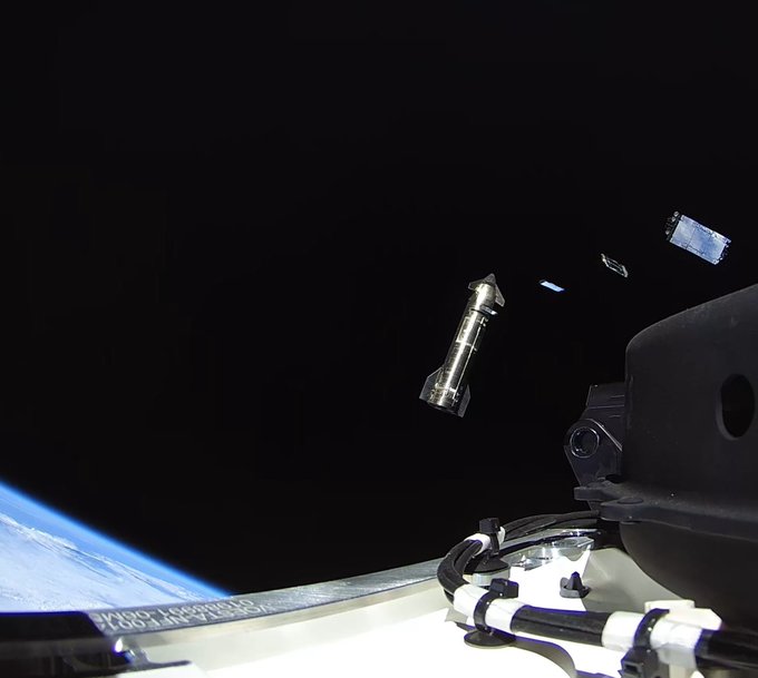 A space photograph showing a cylindrical satellite or rocket stage floating against the blackness of space, with Earth's blue curvature visible in the lower left corner.