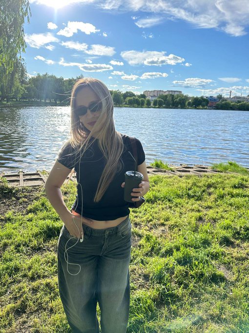 A blonde woman wearing sunglasses and a black crop top stands by a lake holding a coffee cup, wearing earphones, with sun flare in the background.