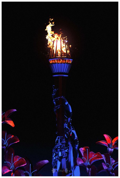 The image features a large, illuminated torch with flames burning brightly at its center, surrounded by flowers of various colors including red, pink, purple, and orange. The flowers are arranged around the base of the torch, creating an eye-catching display against the dark background. The text " Against All Odds" is present in the image, which could be a caption or a description related to the scene.