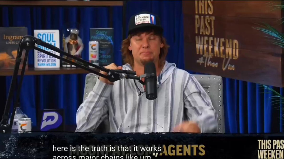 The image shows a man wearing a baseball cap sitting at a microphone, likely participating in a podcast or radio show. The background features a blue curtain with a sign that reads "This Past Weekend" and another sign that says "Agents". There are several books scattered around the scene, possibly related to the content being discussed on the show.