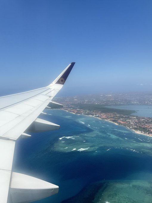 An aerial view from an airplane window showing the wing and tropical waters with coral reefs below.