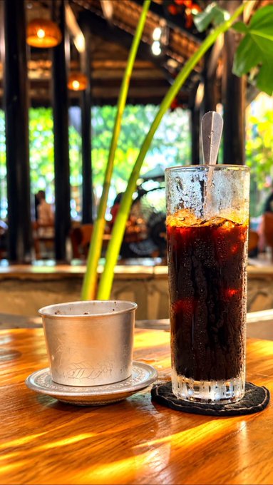 A tall glass of iced dark coffee with a spoon, placed on a wooden table next to a small silver cup and saucer, set in a tropical-themed cafe.