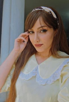 Portrait of a young woman with long brown hair wearing a yellow top with a white ruffled lace collar and a patterned headband, resting her hand near her face.