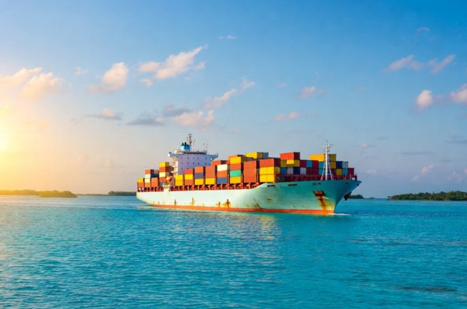 A large cargo ship loaded with colorful shipping containers sails on turquoise water under a blue sky with scattered clouds.