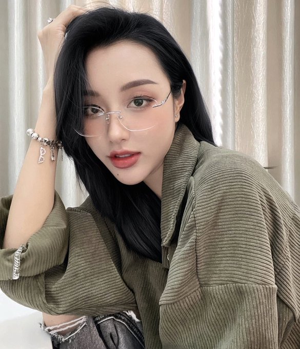 A selfie-style portrait of a young Asian woman with long black hair, glasses, and an olive green ribbed top, wearing a silver charm bracelet.