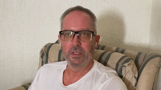 A man with glasses and a short beard wearing a white t-shirt sits on a striped couch in front of a textured white wall.