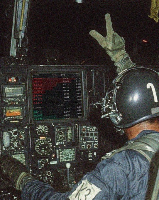 A person in military gear wearing a helmet marked with a '1' sits in an aircraft cockpit making a peace sign with a gloved hand.