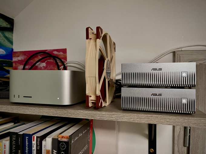 A Mac Mini, a Noctua fan, and two ASUS external drives sit on a wooden shelf above a collection of books including a Trauma manual.