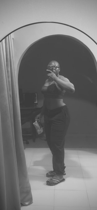 A black-and-white mirror selfie of a woman wearing sunglasses, a bra top, and baggy pants, holding a snack package in a room with an arched doorway and curtain.