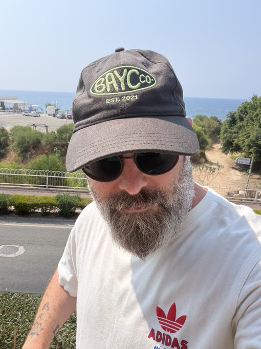 A selfie of a bearded man wearing sunglasses and a black "BAYC co. EST. 2021" cap, dressed in a white Adidas t-shirt, with a road, trees, and ocean visible in the background.